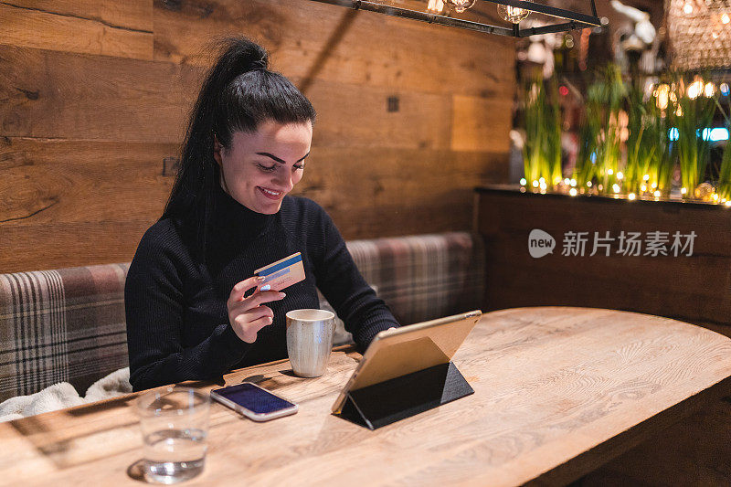 Beautiful young woman holding her credit card while using a tablet in a café